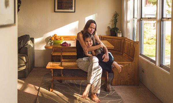 Mom sitting at kitchen table hugging her young daughter.